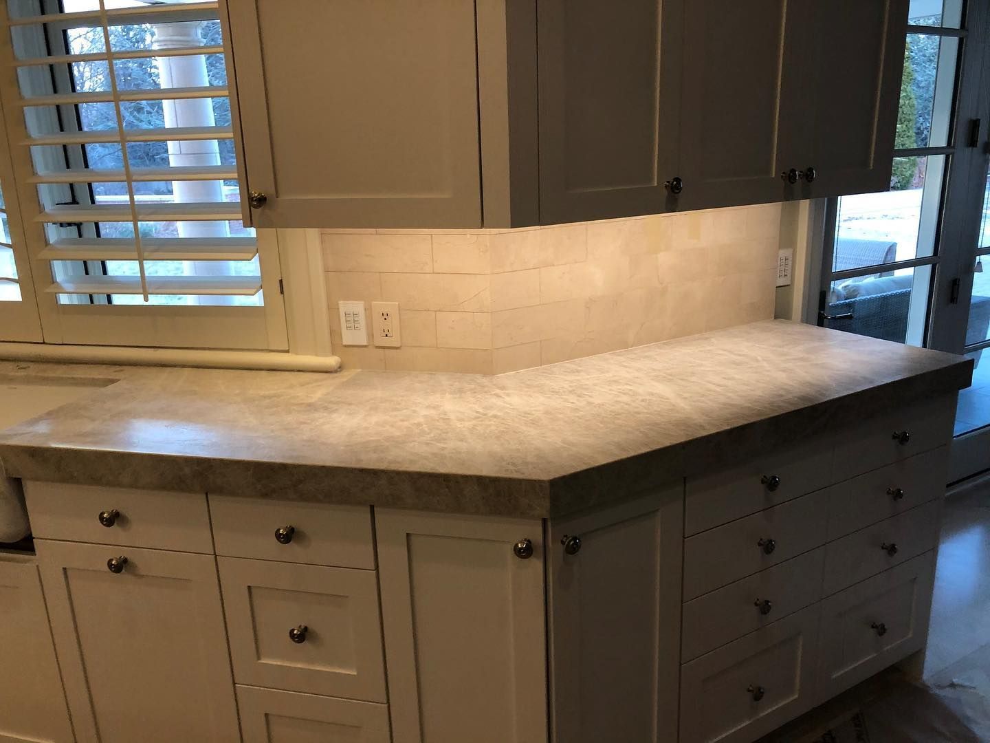 Kitchen with light-colored cabinets, countertop, and backsplash. A window with shutters is on the left.