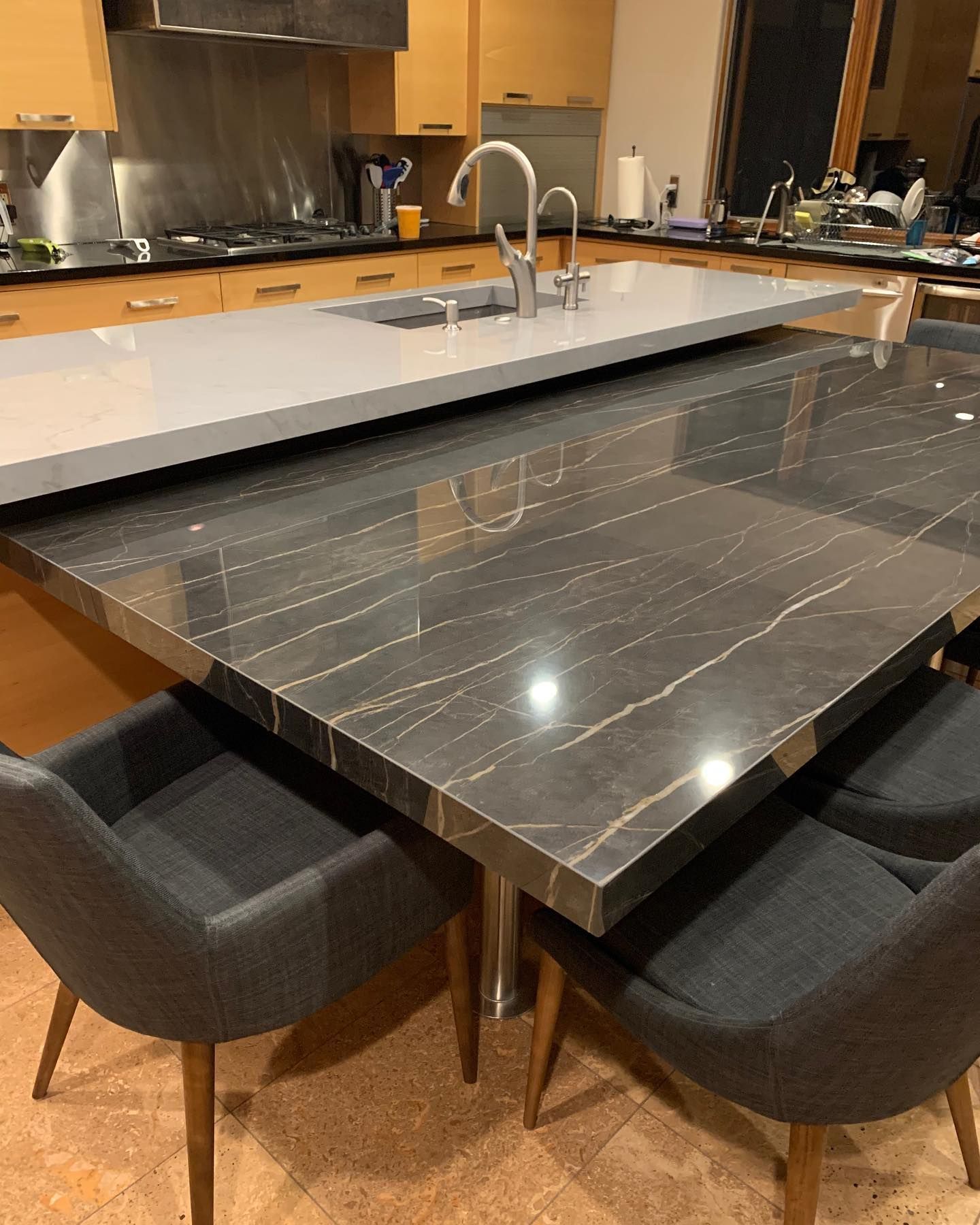 Kitchen island with dark marble countertop, gray chairs, and a light-colored countertop with sink.