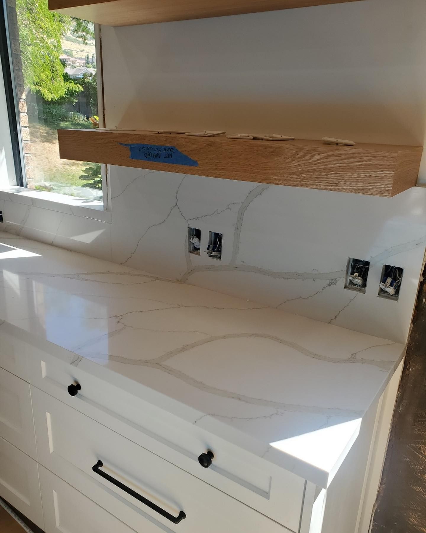 White countertop and backsplash with marble-like veining, beneath a wooden shelf, above white cabinets.