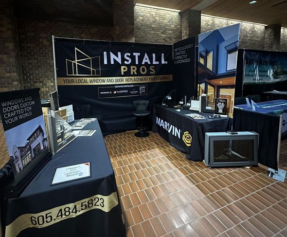 Trade show booth with black tablecloths and banners, showcasing "Install Pros" with their logo and phone number.