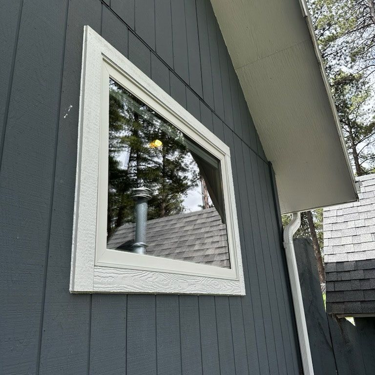 A square window with a white frame on a gray house exterior. The window reflects the surroundings.