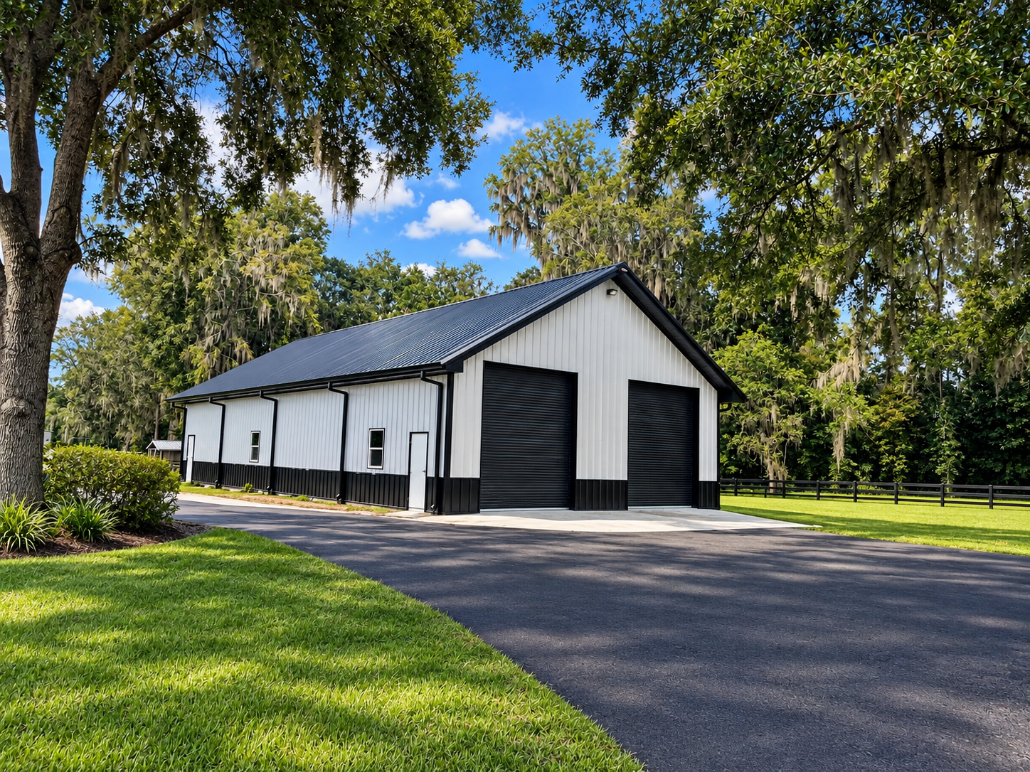 White barn with black doors along a paved drive, framed by green lawn and trees under a blue sky