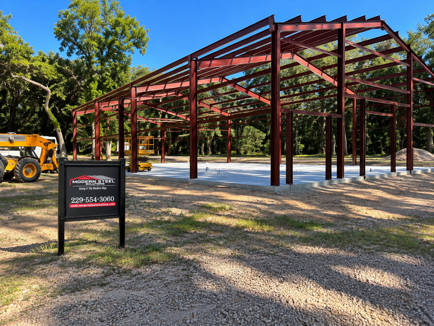 Partially built steel frame structure on gravel, with a yellow loader and trees in the background