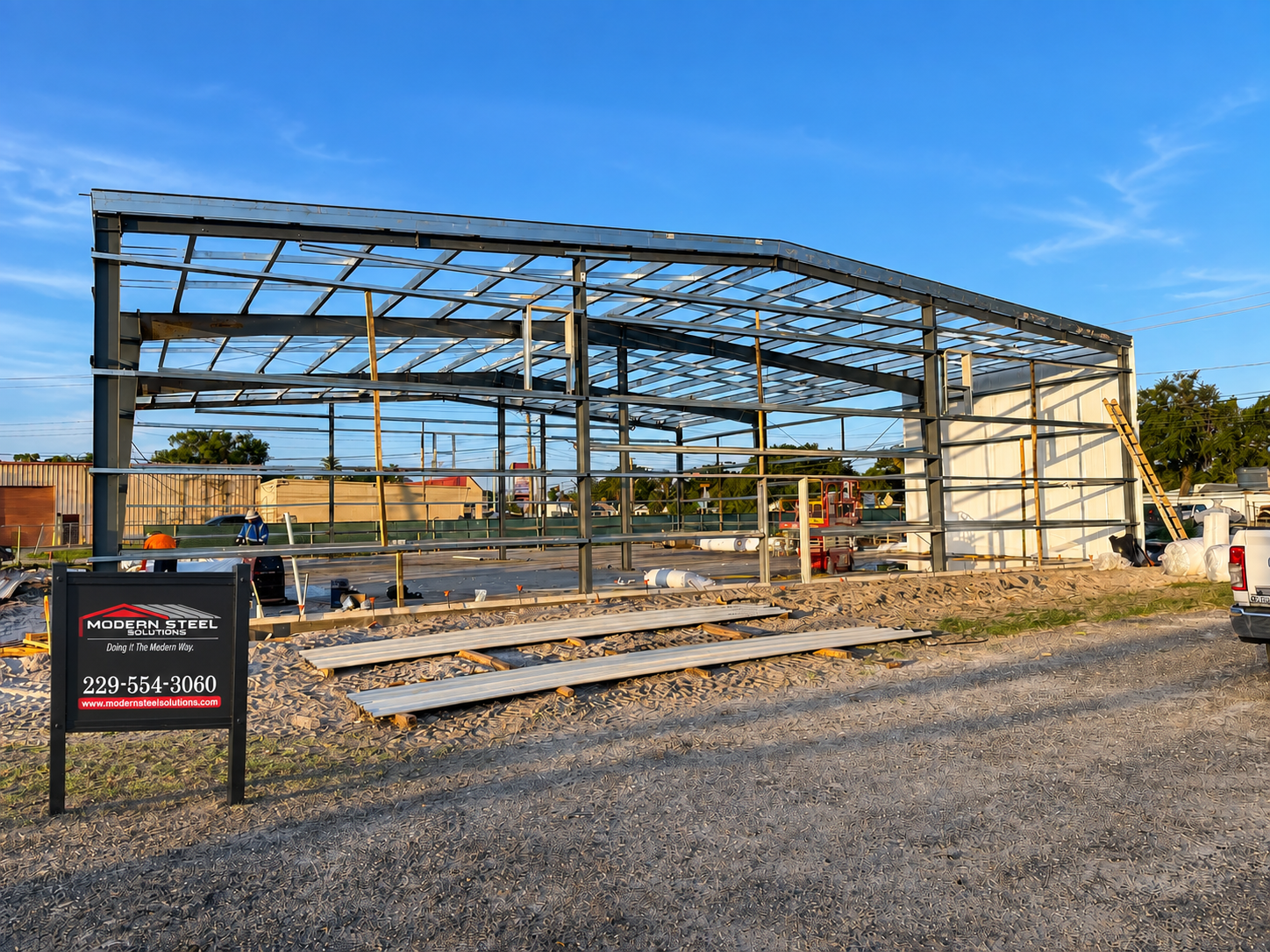 Steel building frame under construction on a gravel lot with scaffolding and a blue sky.