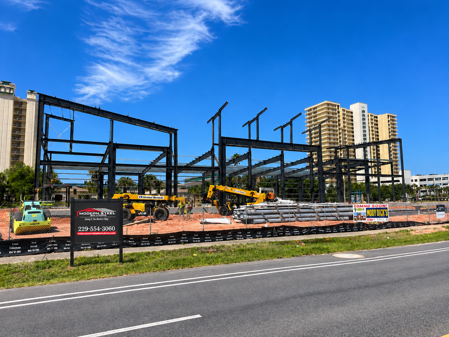 Construction site with steel framework and yellow machinery beside a roadway under a blue sky