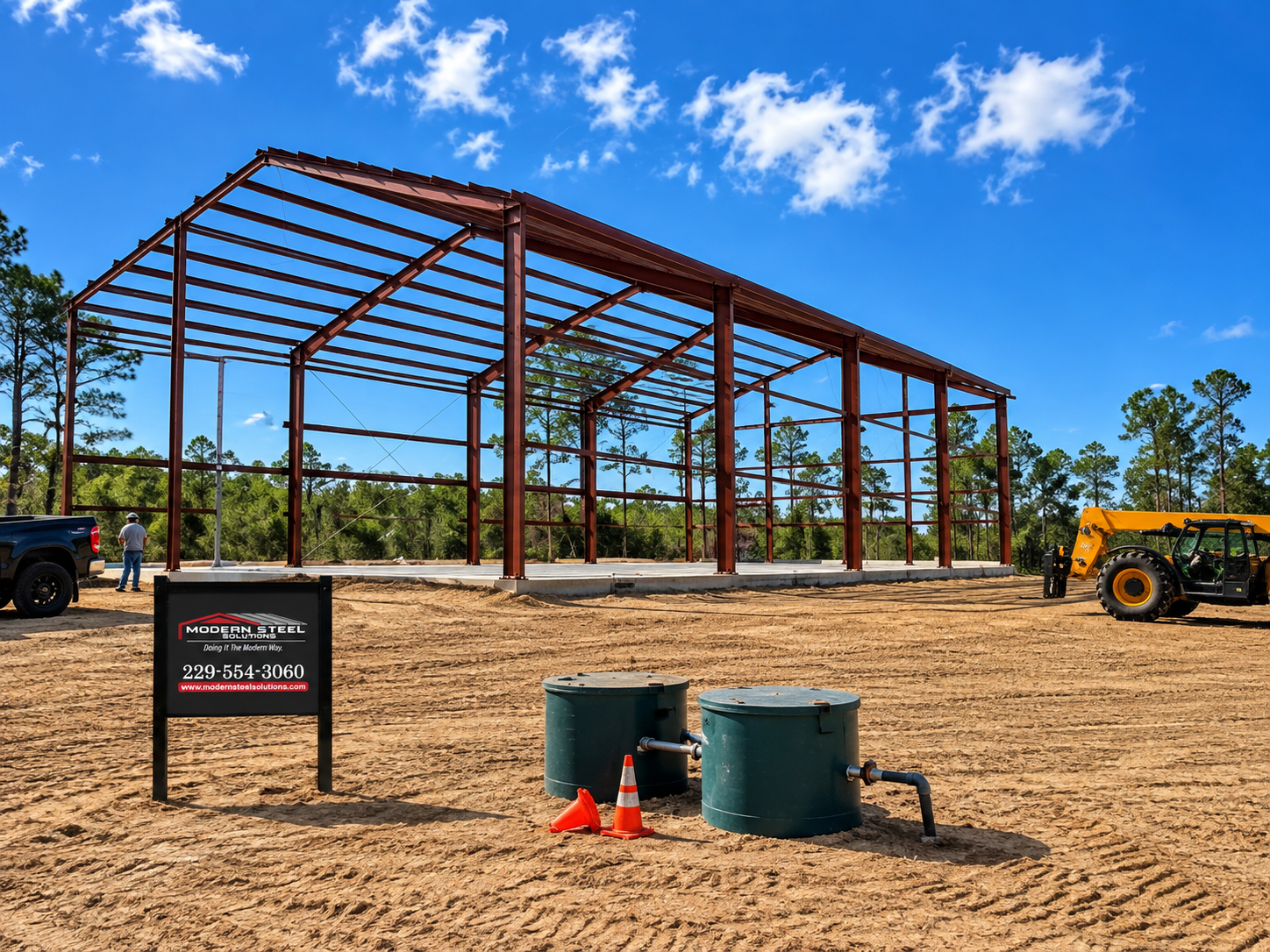 Steel-frame building under construction on a dirt site, with construction vehicles and blue sky.