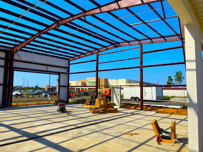 Steel building frame under construction with blue roof panels and equipment on a concrete slab