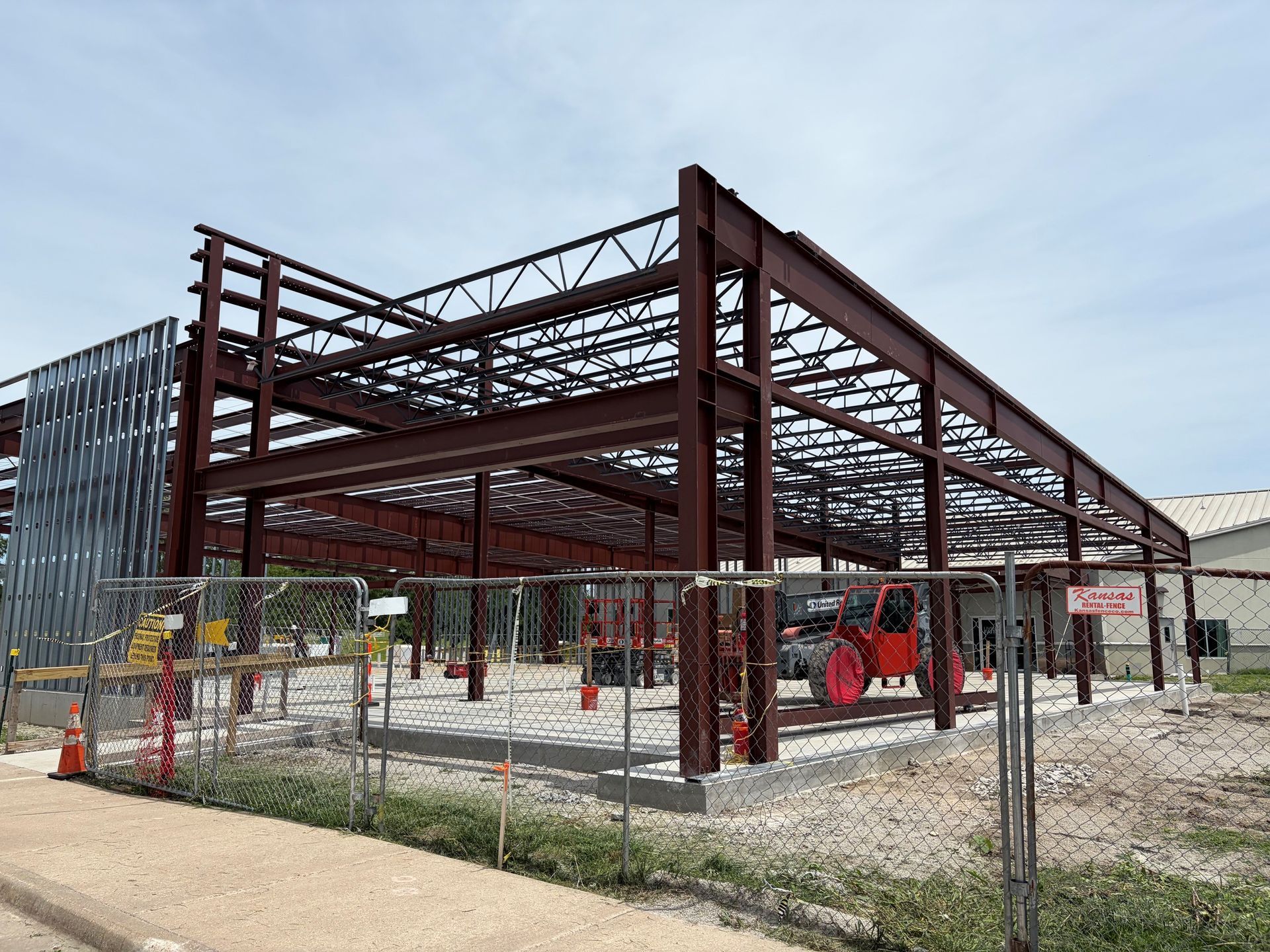 Steel building frame under construction with workers, fencing, and equipment on a gravel lot.