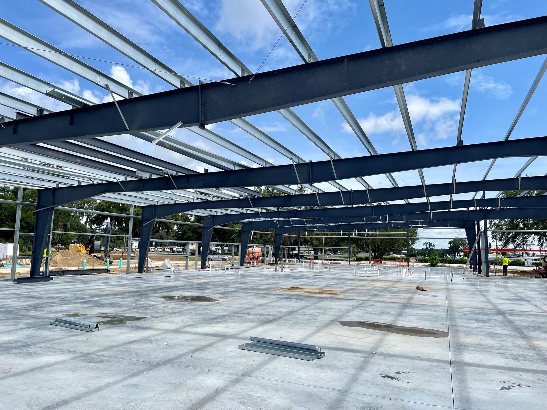 Steel-frame building under construction with a concrete floor and open roof against a blue sky.
