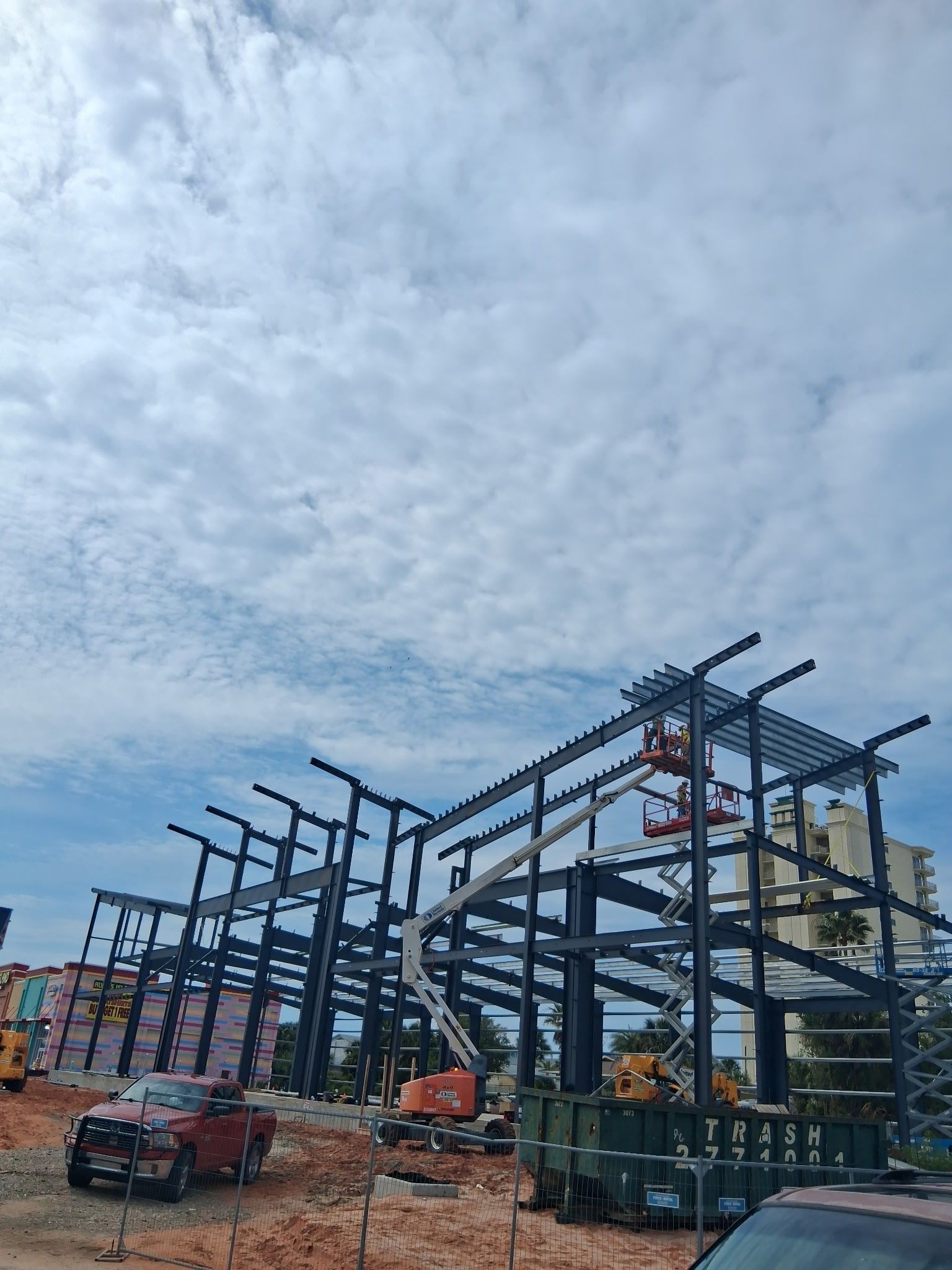 Steel building frame under construction on a cloudy day, with cranes and trucks on site.