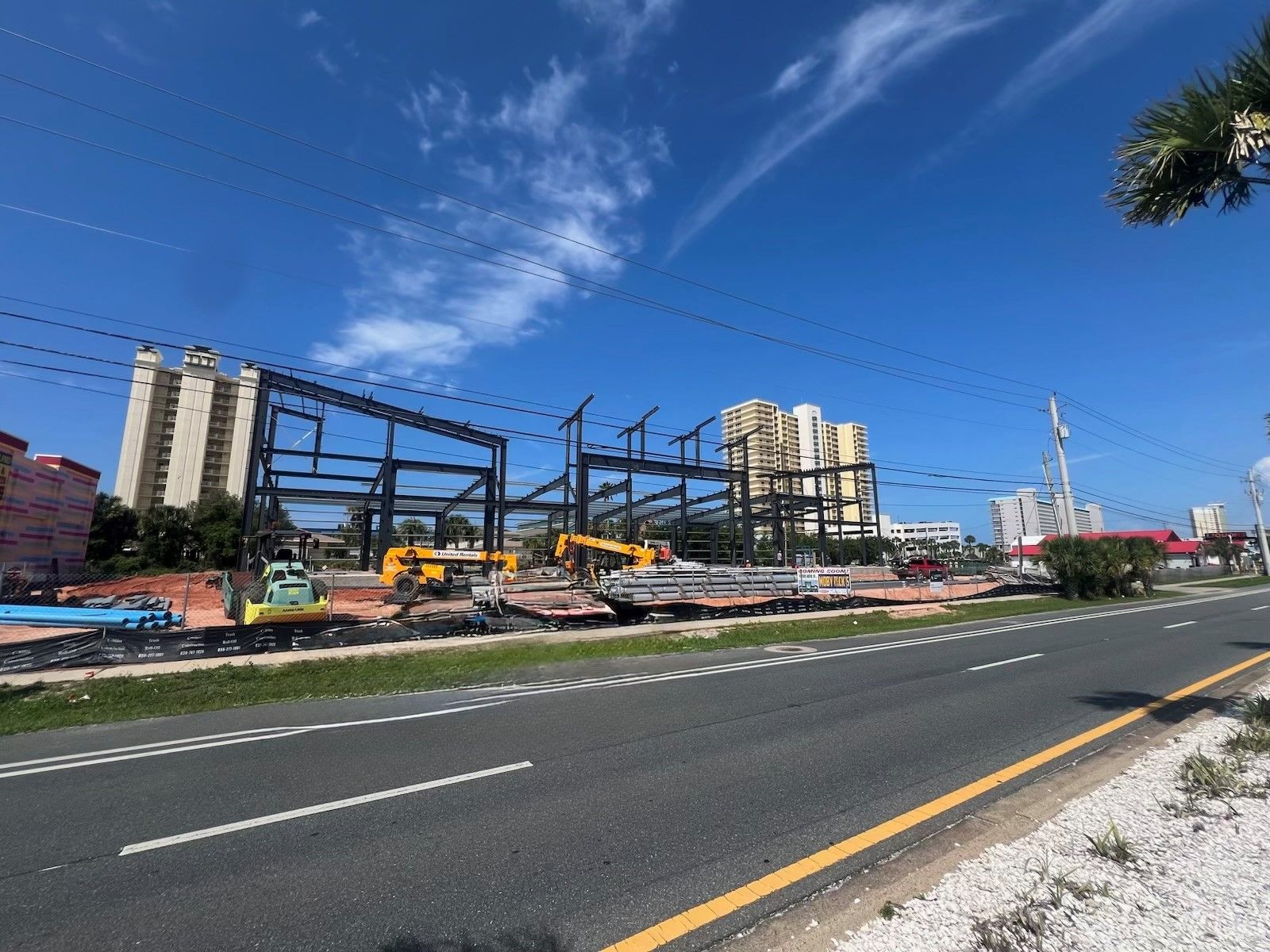 Construction site beside a wide road under a bright blue sky, with cranes and partially built steel structures.