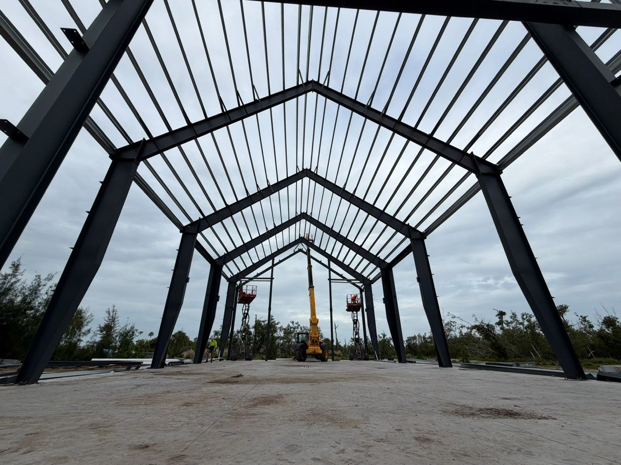 Steel frame pavilion under construction on a cloudy day, with a crane in the distance and gravel ground.