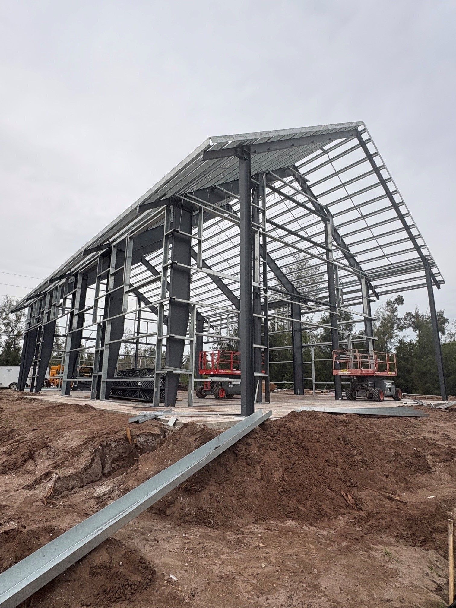 Steel-frame building under construction on a dirt site, with roof trusses and workers visible in the distance.