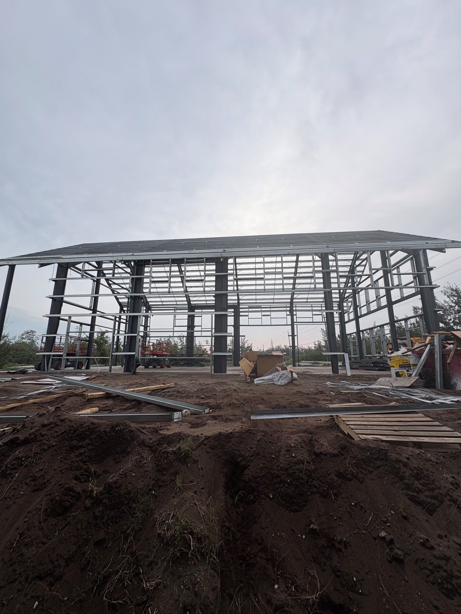 Steel frame of a building under construction on a muddy site, with equipment and workers in the background.