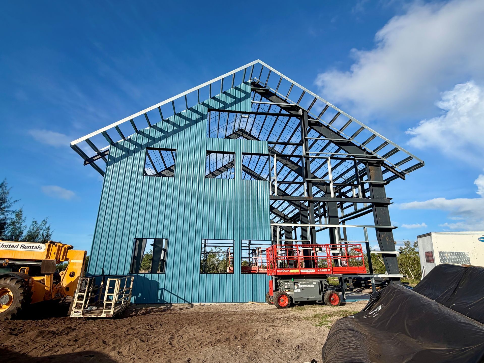 Blue building under construction with exposed steel framing, heavy machinery, and piles of gravel under a blue sky.