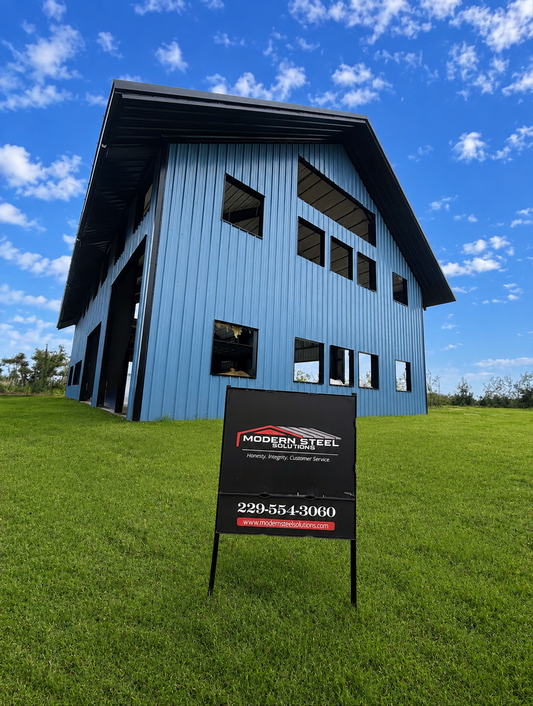 Blue modern house on a grassy hill under a bright sky, with a black real estate sign in front.