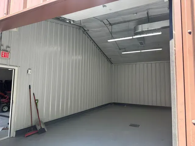Empty metal storage unit with open doorway, white corrugated walls, gray floor, and cleaning tools by the door.