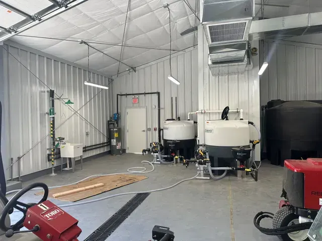 Industrial workshop with white metal walls, large equipment tanks, hoses, and a red machine on the floor.