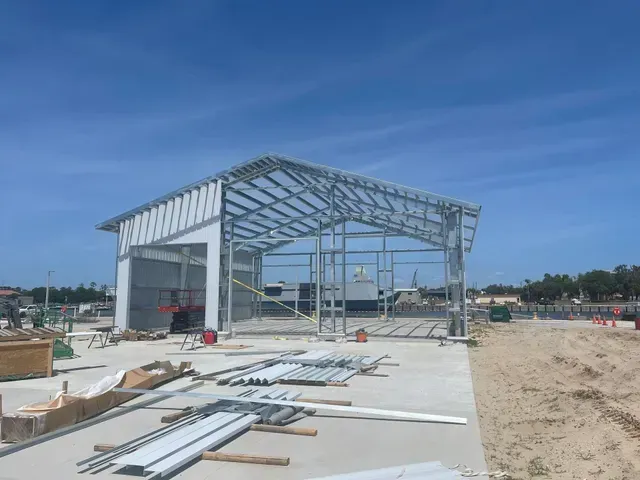 Steel-framed building under construction on a sandy site with materials and clear blue sky.