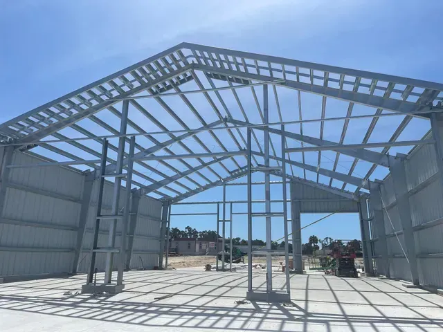 Steel-framed building under construction with a gabled roof skeleton against a blue sky.