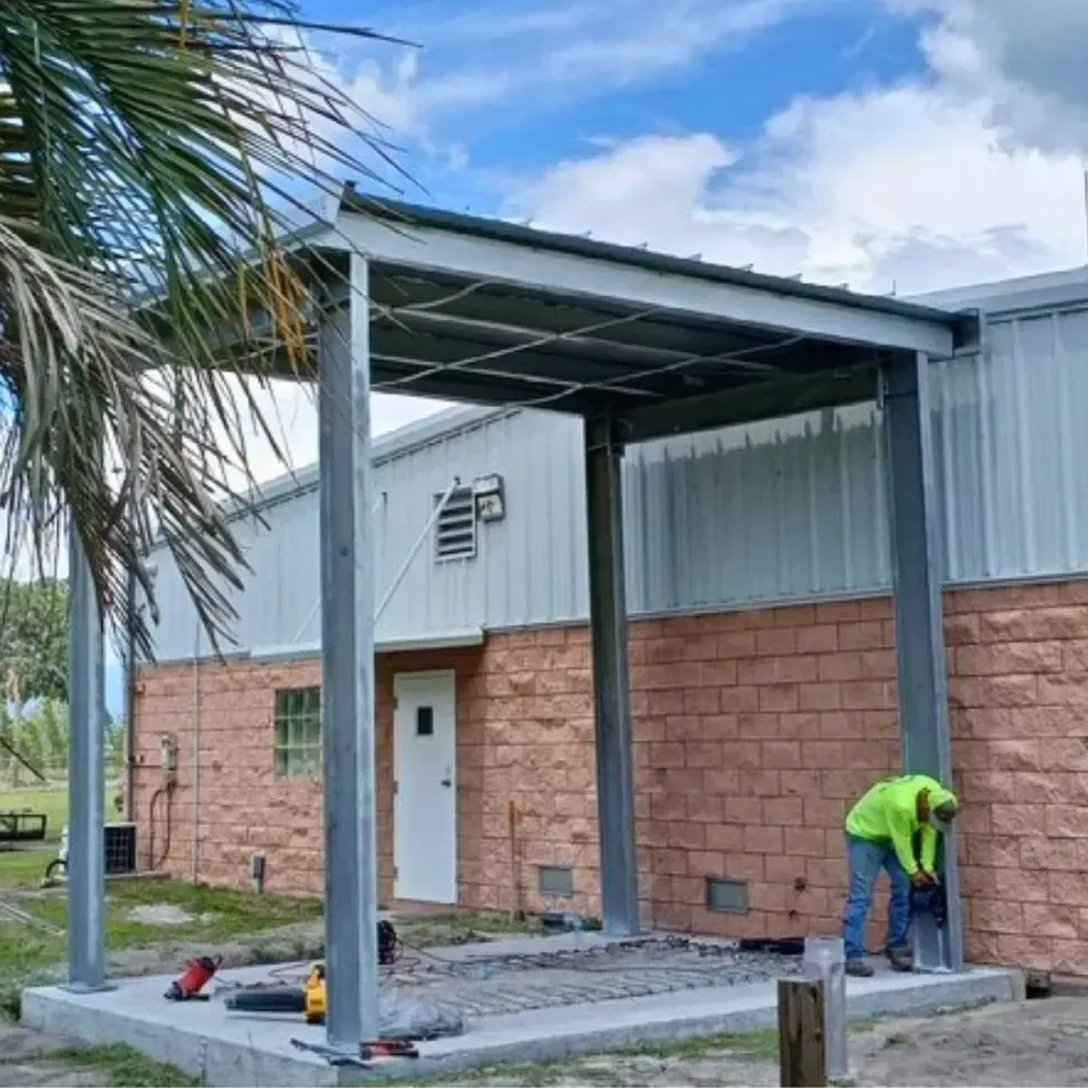 Two workers building a covered patio beside a brick-and-metal building, with one drilling near the post base.
