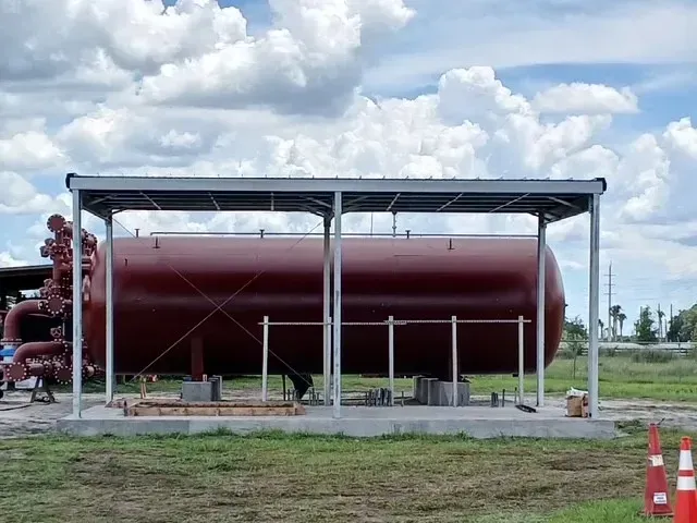 Large red industrial tank under a metal canopy in a grassy field, with pipes and orange cones nearby