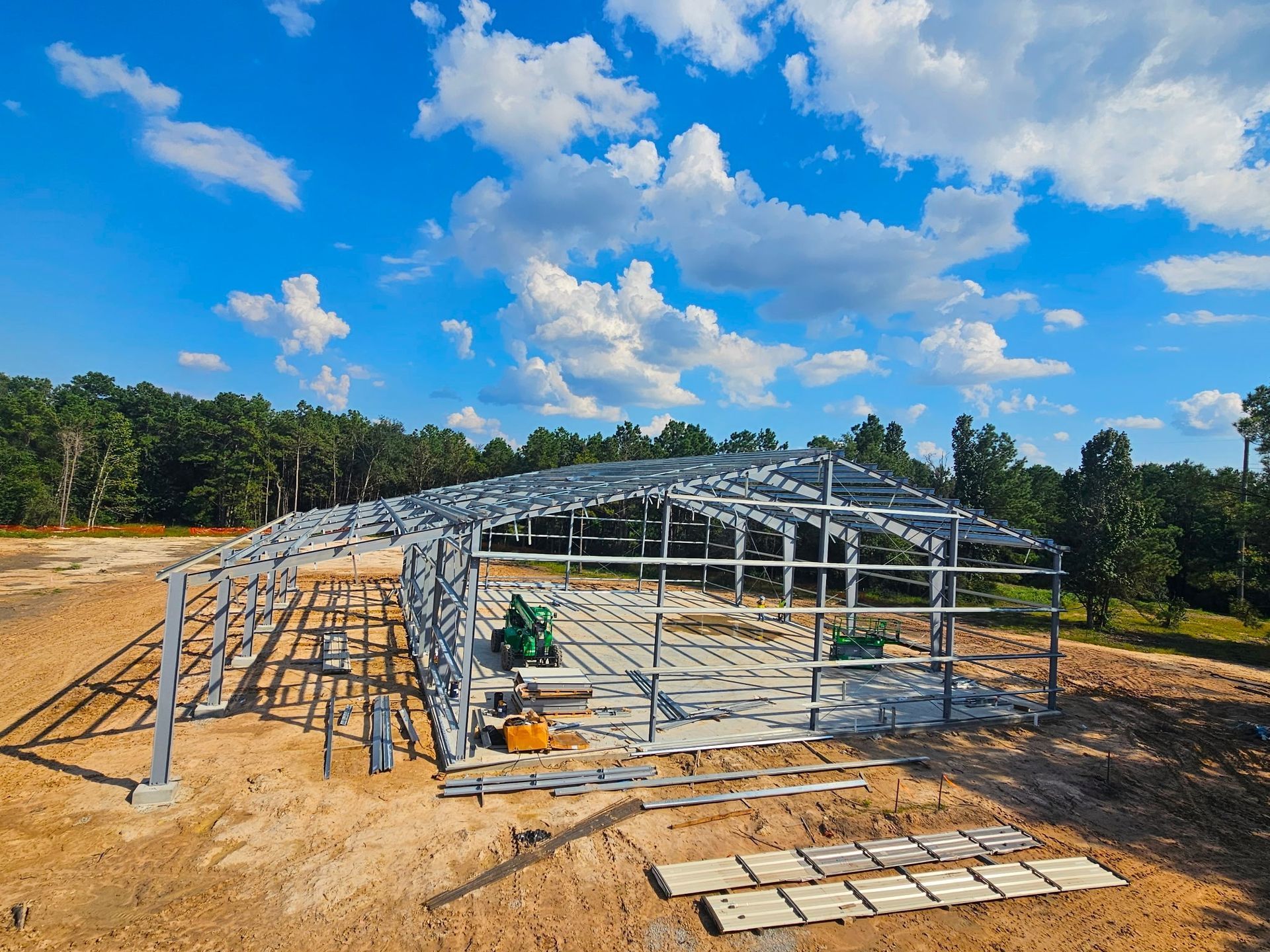 Steel-frame building under construction on a dirt lot, with blue sky and clouds overhead