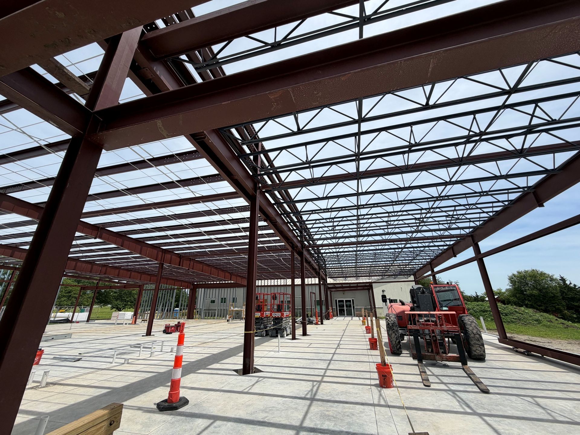 Steel-frame building construction site with a red lift, orange cones, and a concrete slab under a truss roof