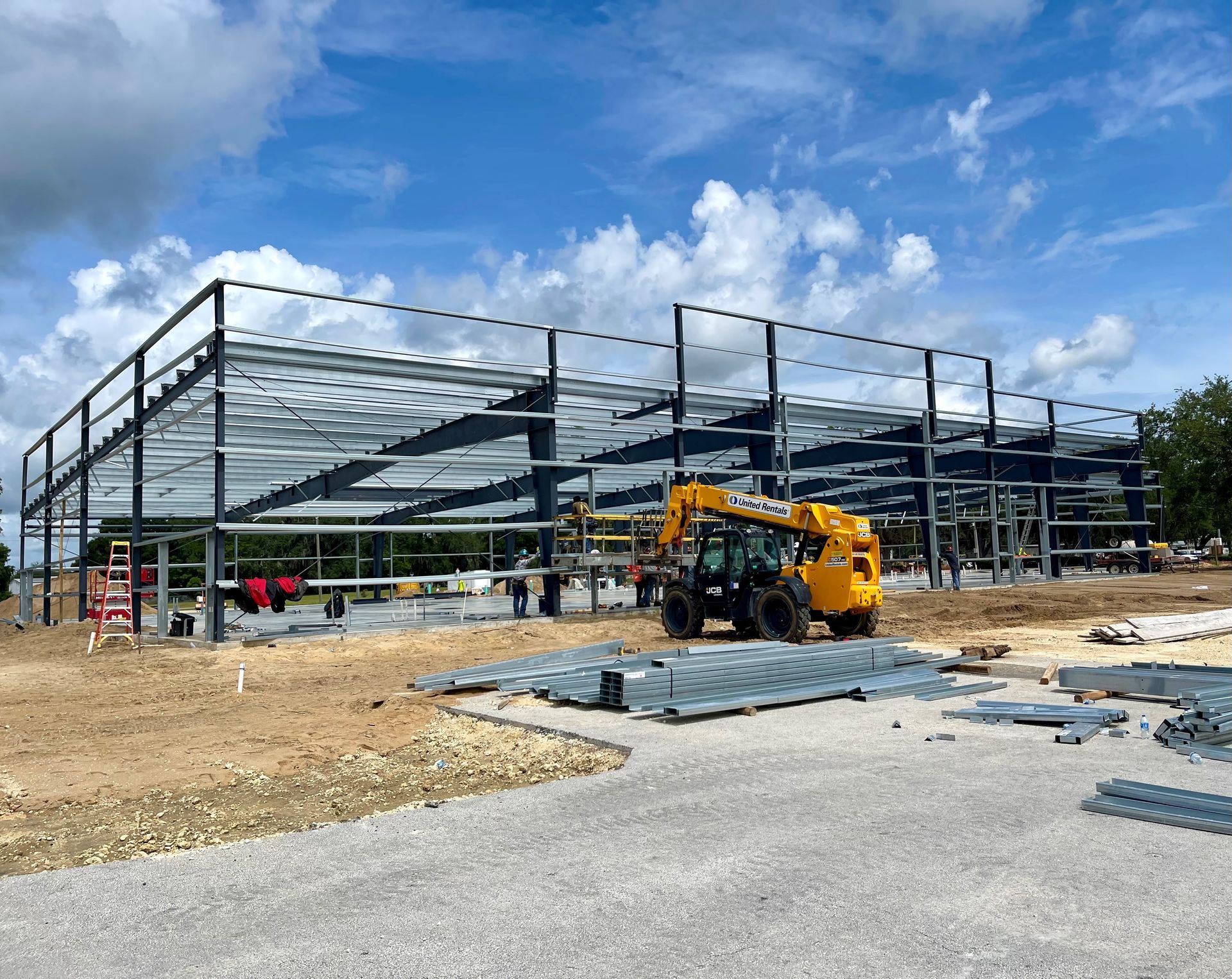 Steel-frame building under construction with a yellow loader, dirt lot, and blue sky.
