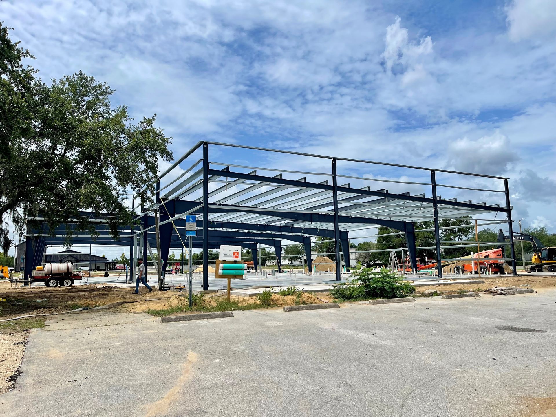 Steel frame of a building under construction beside a paved lot, with trees and equipment in the background