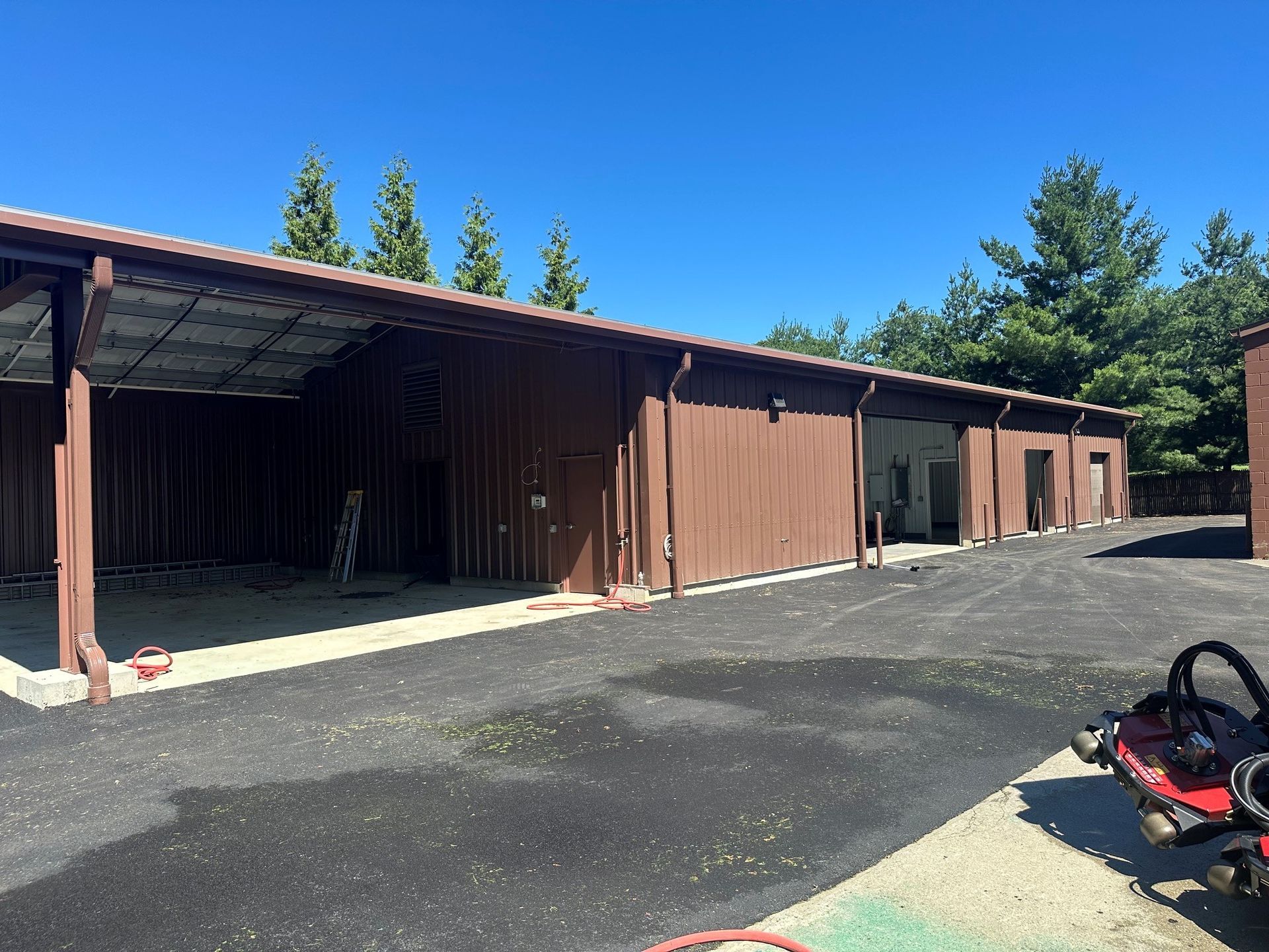 Long brown barn with open bays beside a paved lot, under a bright blue sky and tall trees