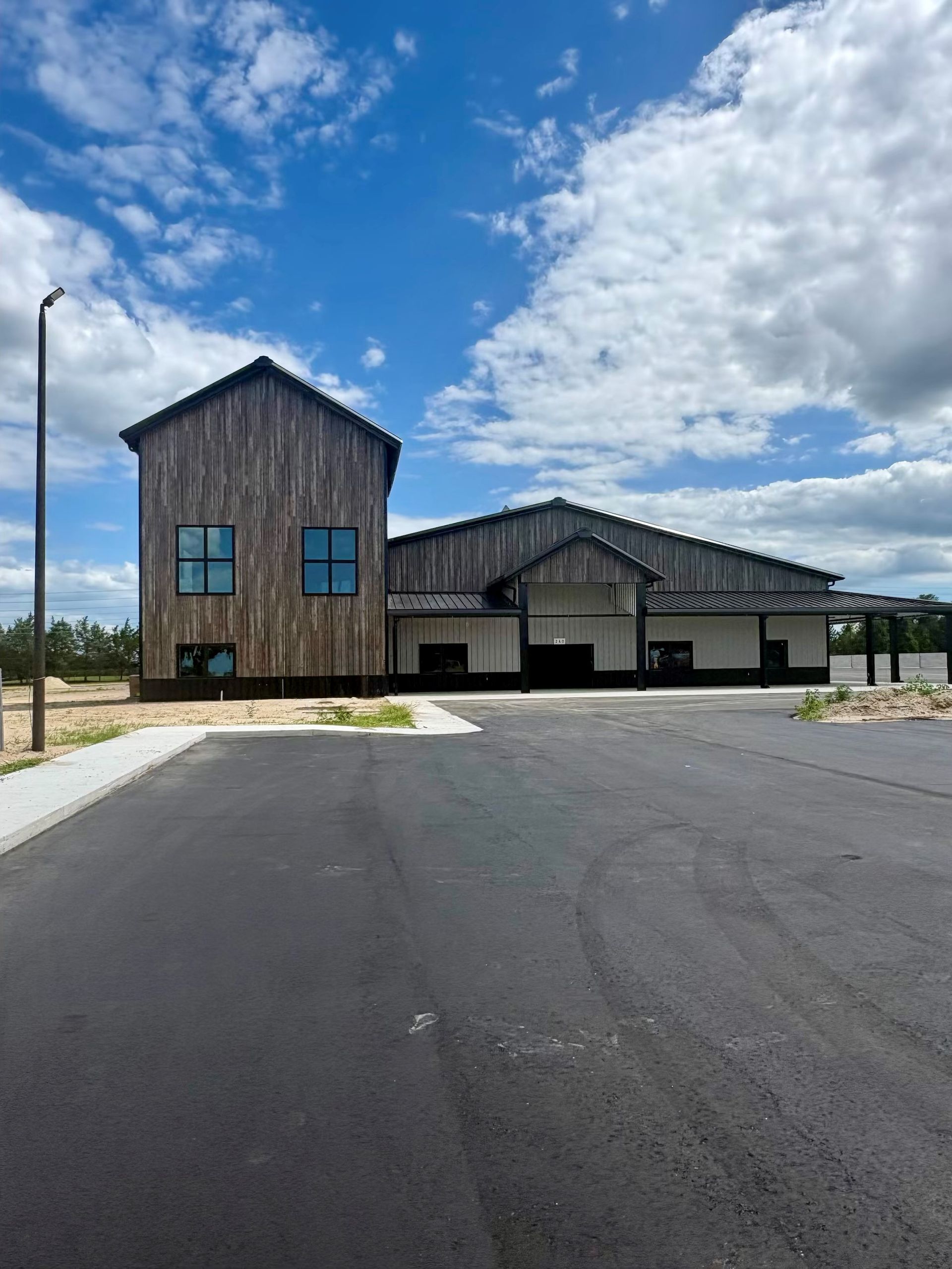 New wooden building with blue windows and a large paved parking lot under a partly cloudy sky