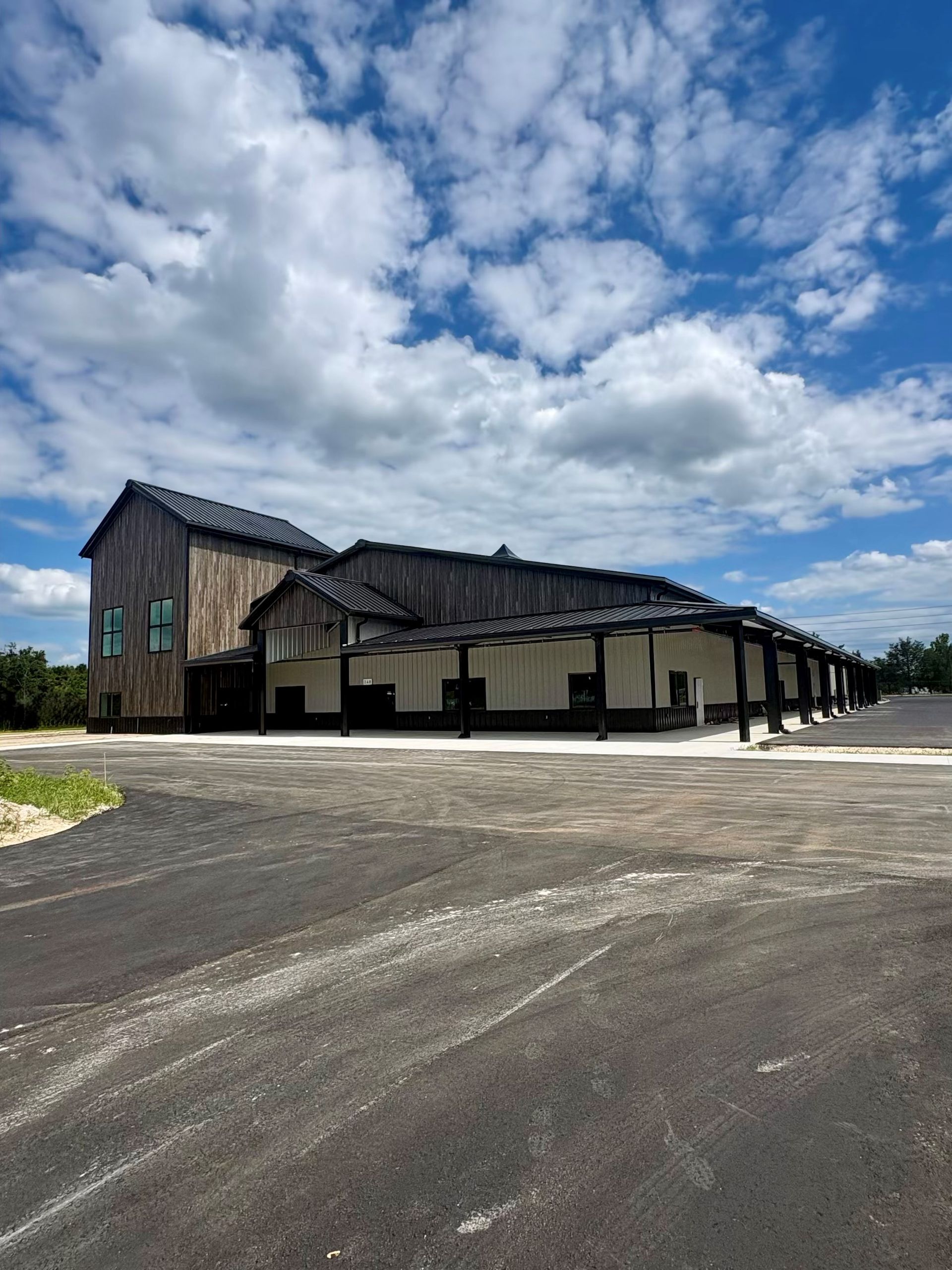 Large rustic building with dark roof beside an open paved lot under a partly cloudy sky