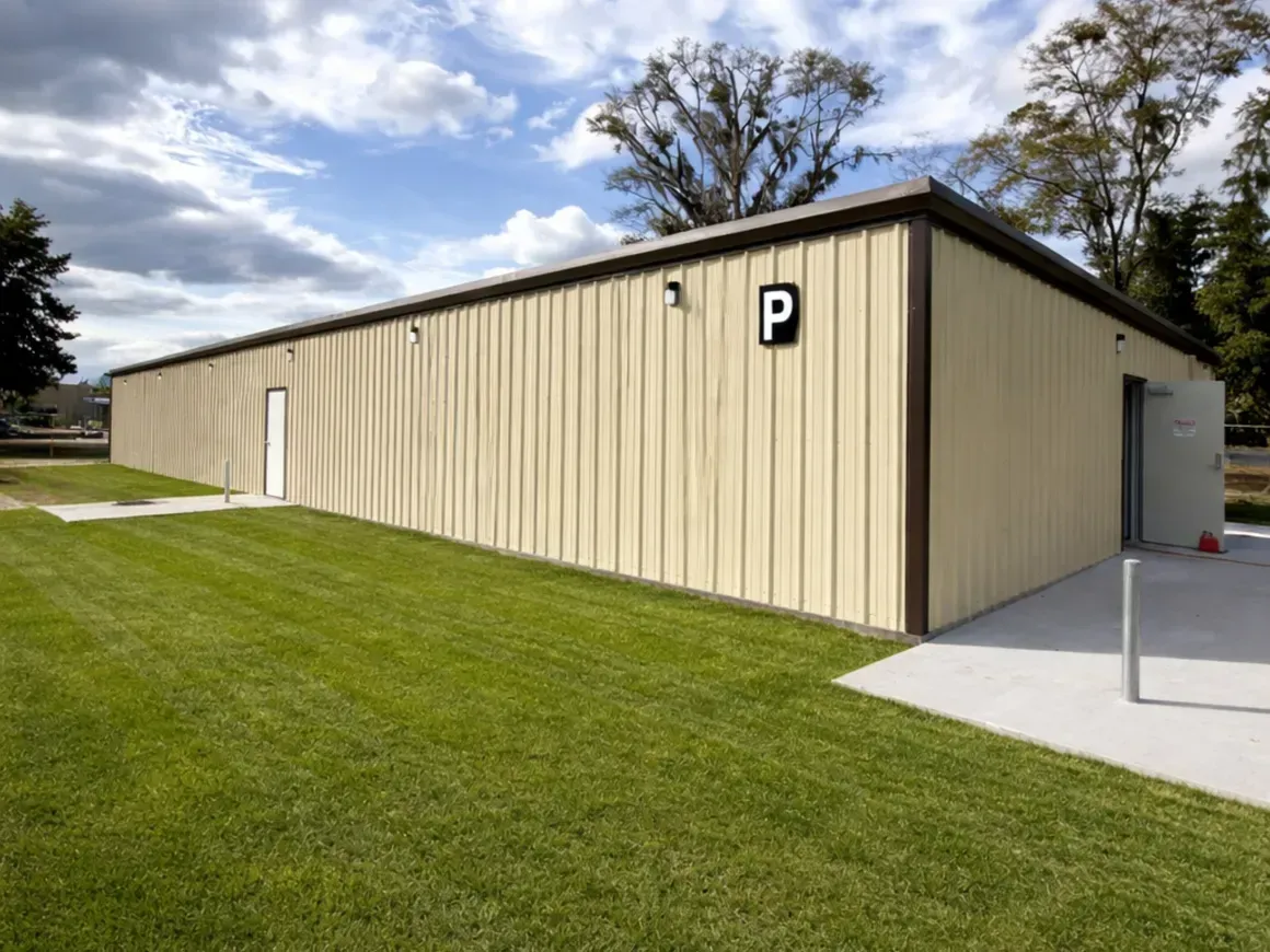 Beige metal restroom building beside a grassy lawn under a partly cloudy sky