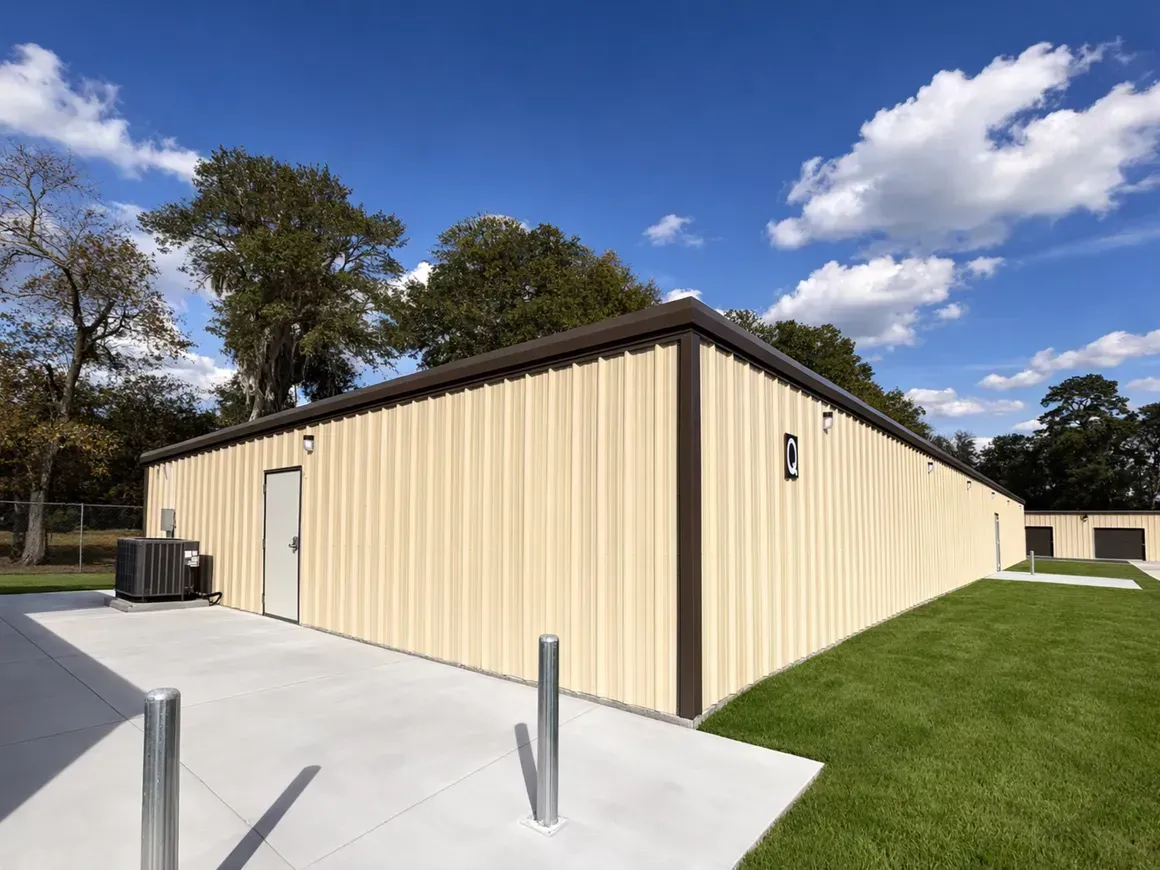 Beige metal building with a concrete pad, grassy lawn, and blue sky with clouds