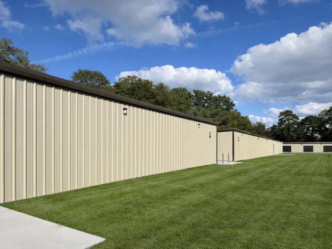 Beige storage buildings beside a green lawn under a blue sky with clouds