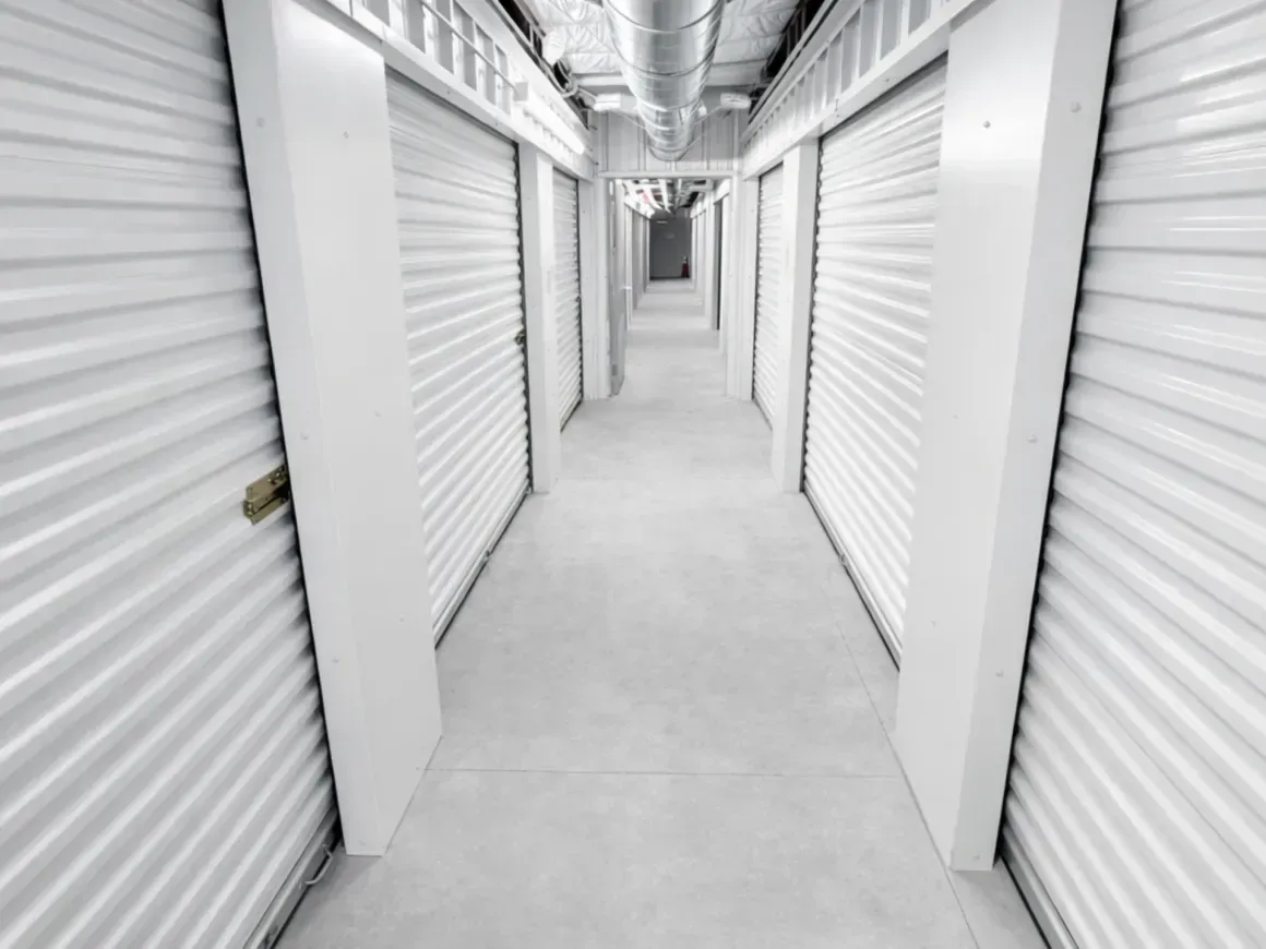 White storage unit hallway with closed roll-up doors on both sides, viewed in perspective