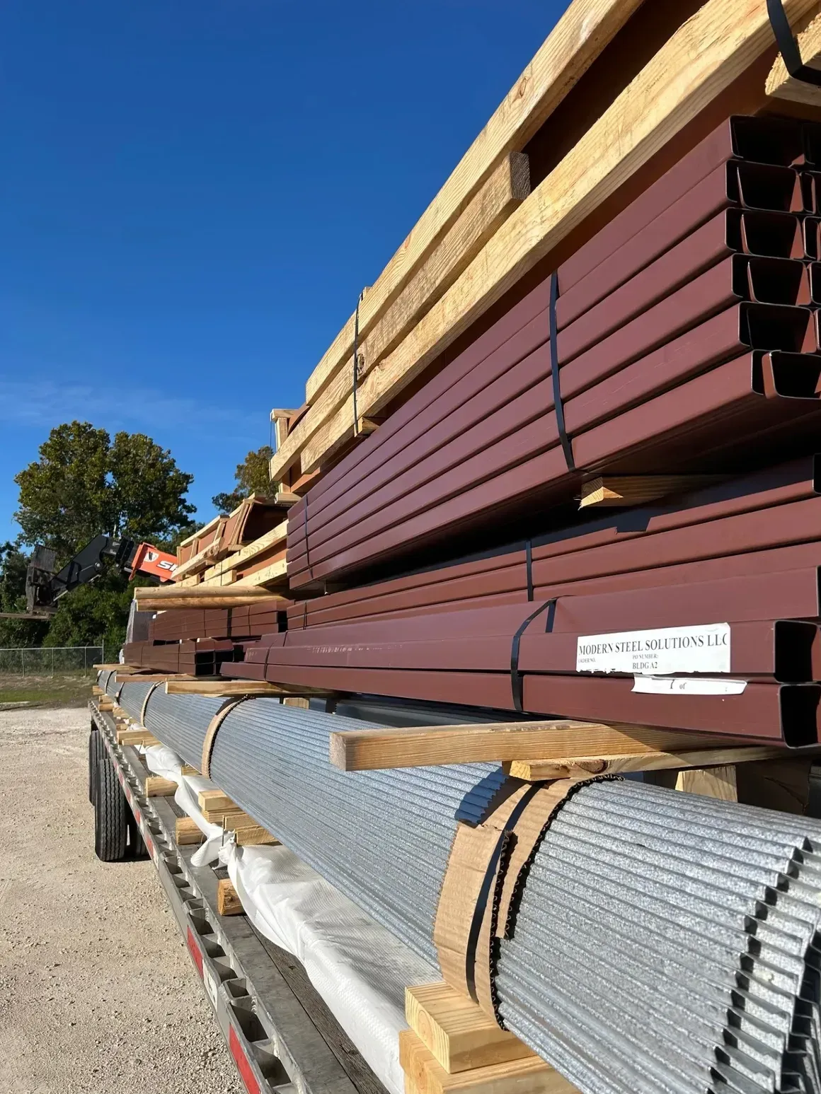 Stacked metal beams and lumber secured on a flatbed trailer outdoors under a clear blue sky.
