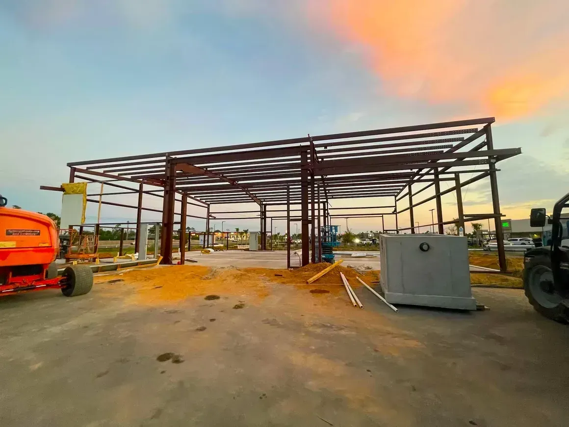 Steel frame building under construction at sunset, with equipment and materials on site.