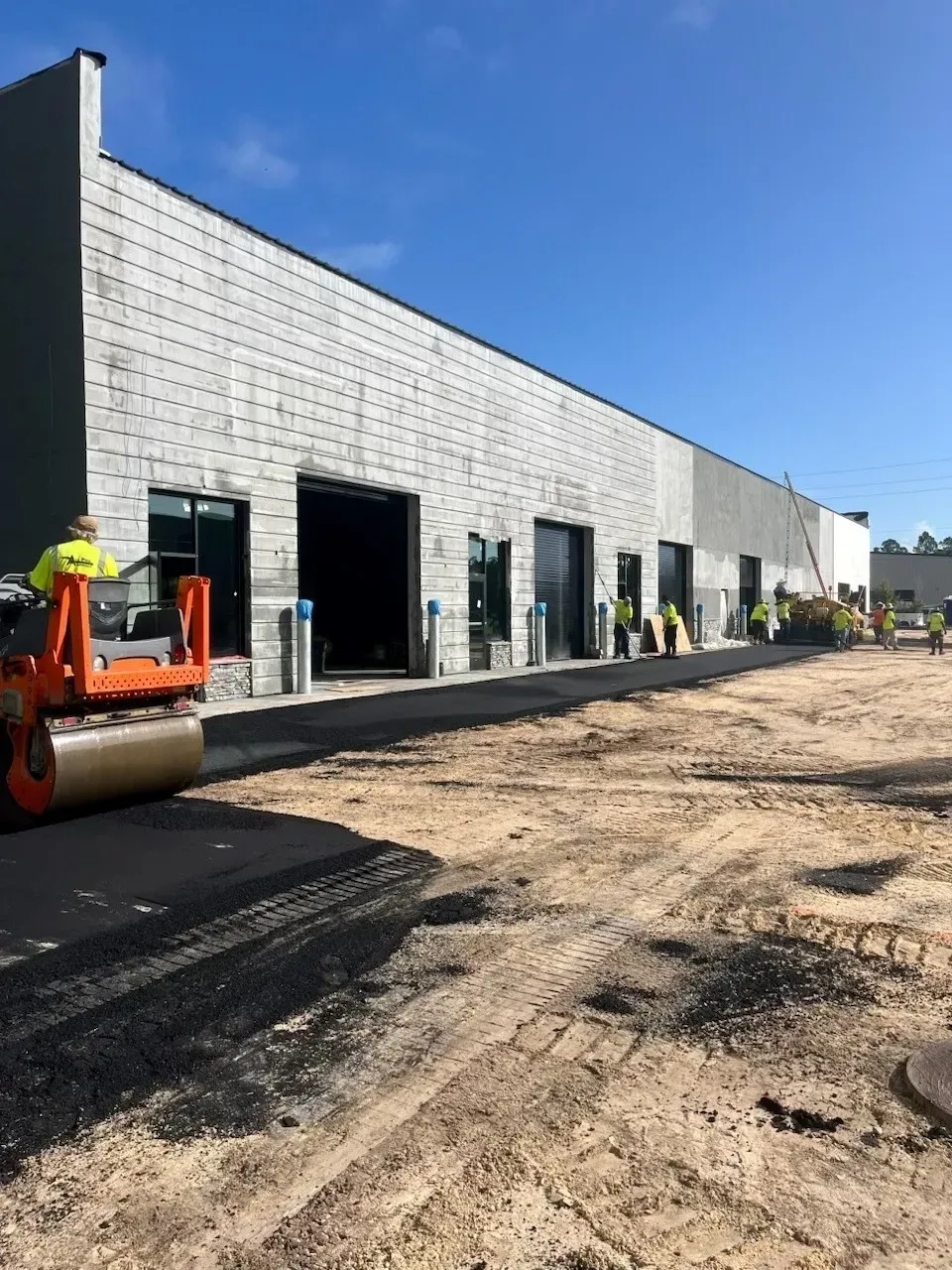 Construction site with a long concrete building, open doorways, and workers near an orange roller under a blue sky