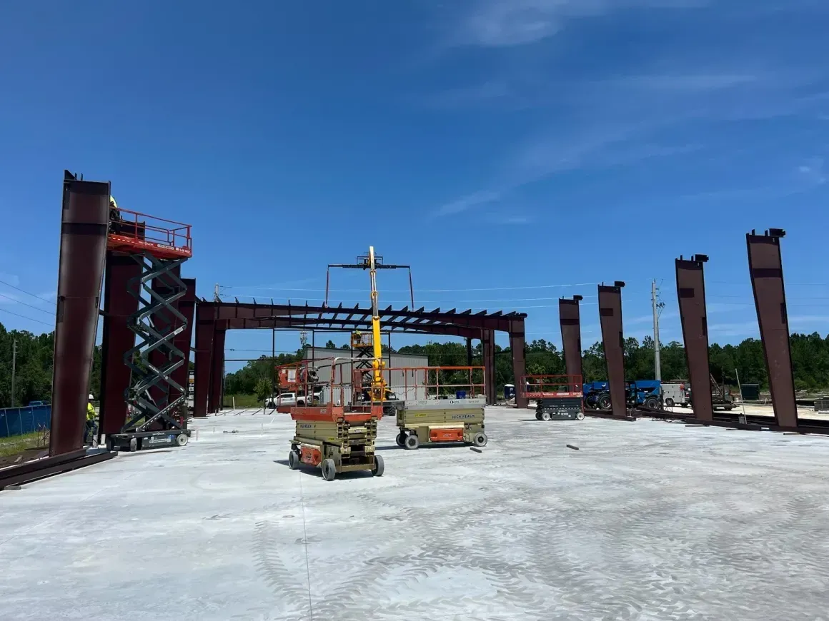 Construction site with steel frame columns, crane, and equipment on a concrete slab under a blue sky