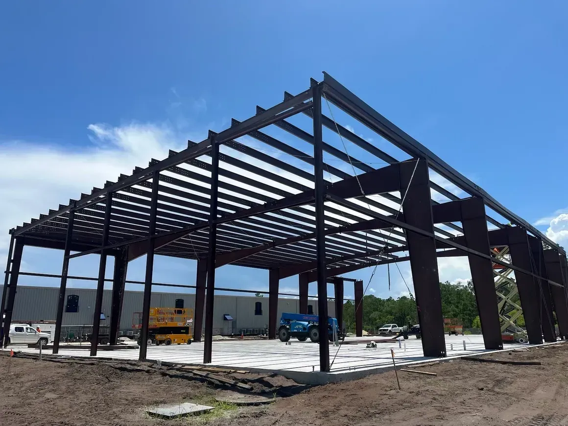 Steel warehouse frame under construction on a concrete slab, with construction vehicles and a blue sky.