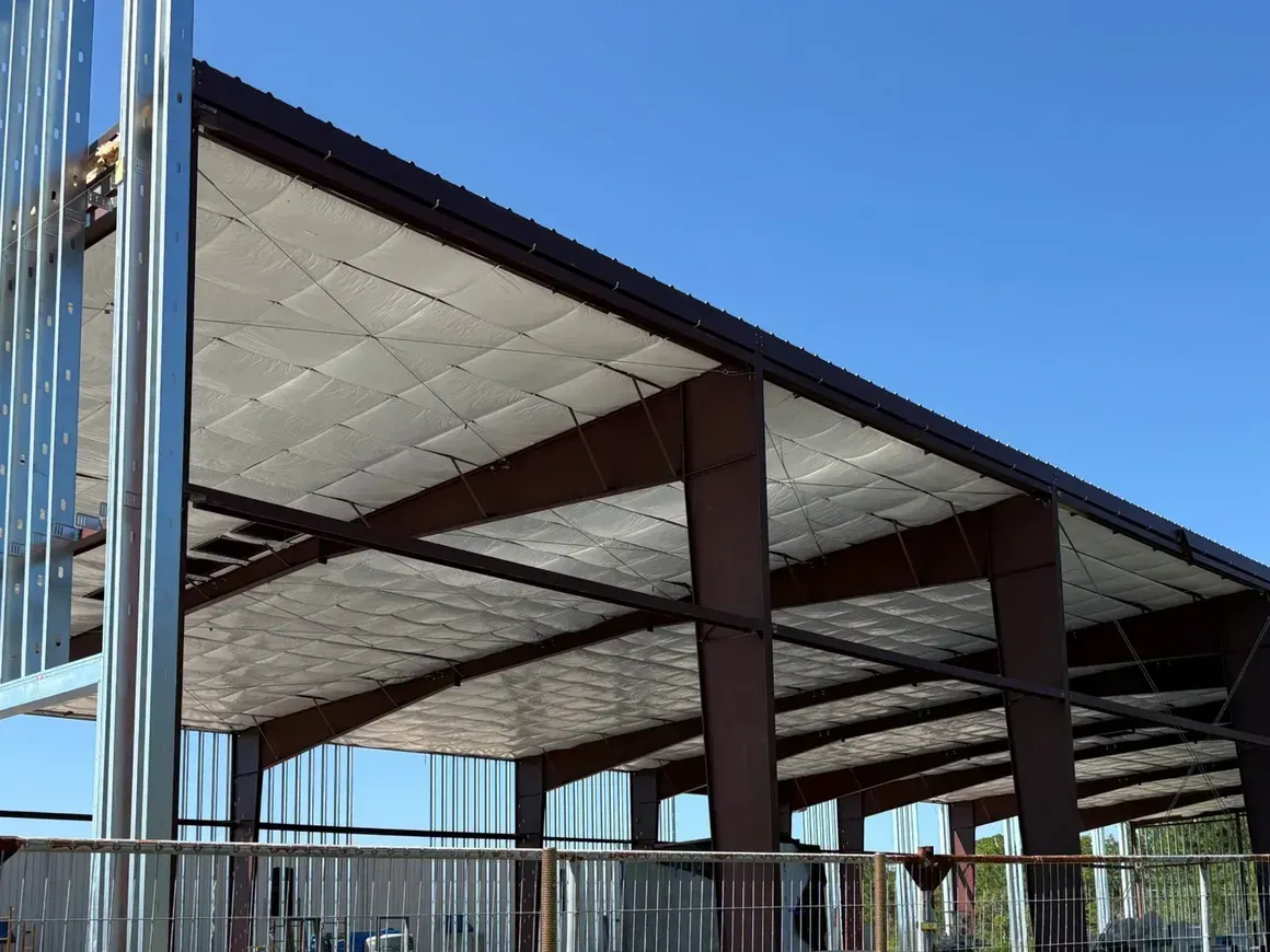 Steel building frame with dark beams and a white insulated roof under a blue sky