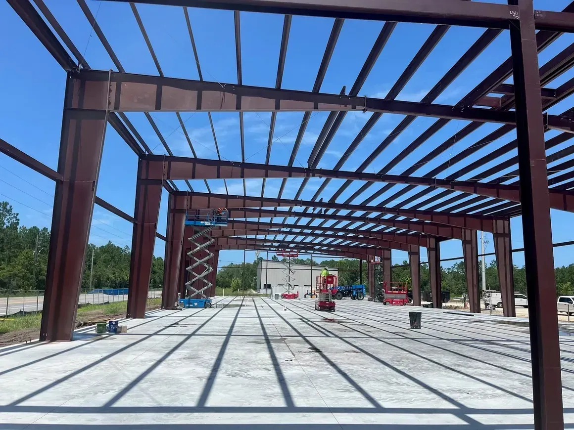 Steel frame building under construction on a sunny day, with concrete floor and red machinery in the distance