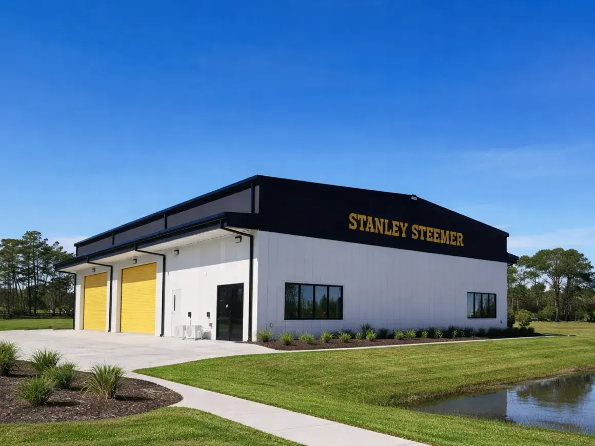 Modern white building with black roof and yellow doors labeled “Stanley Steer” beside a pond
