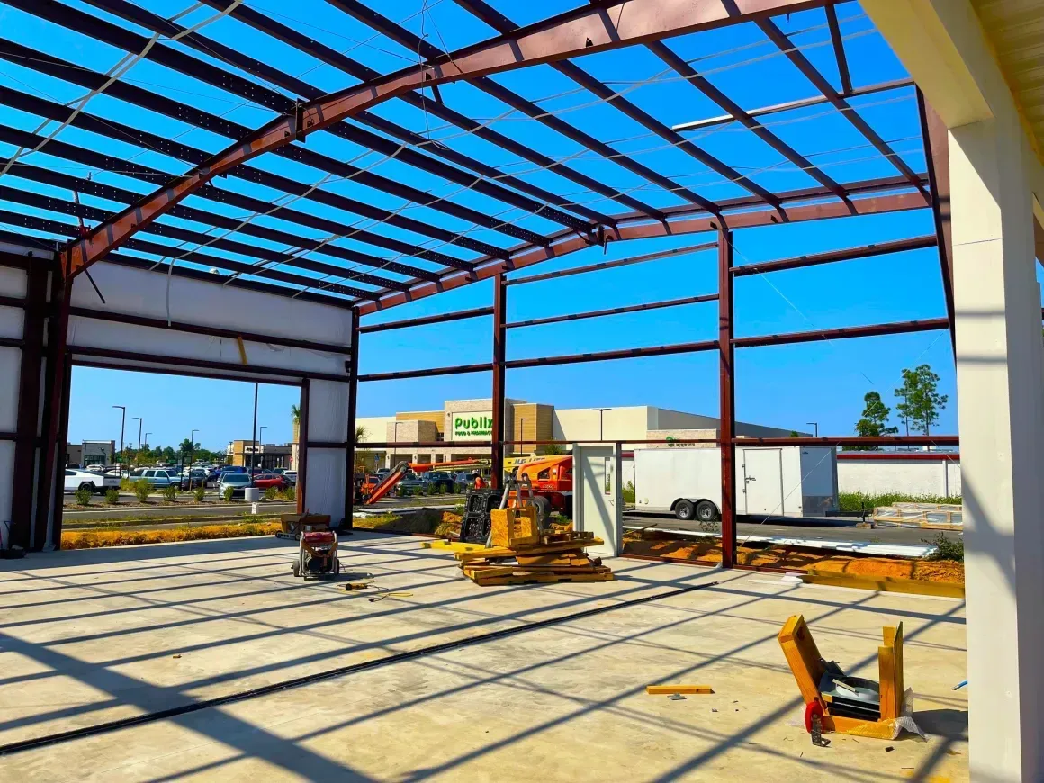 Steel-frame building under construction with yellow equipment and sunlight casting shadows on the concrete floor