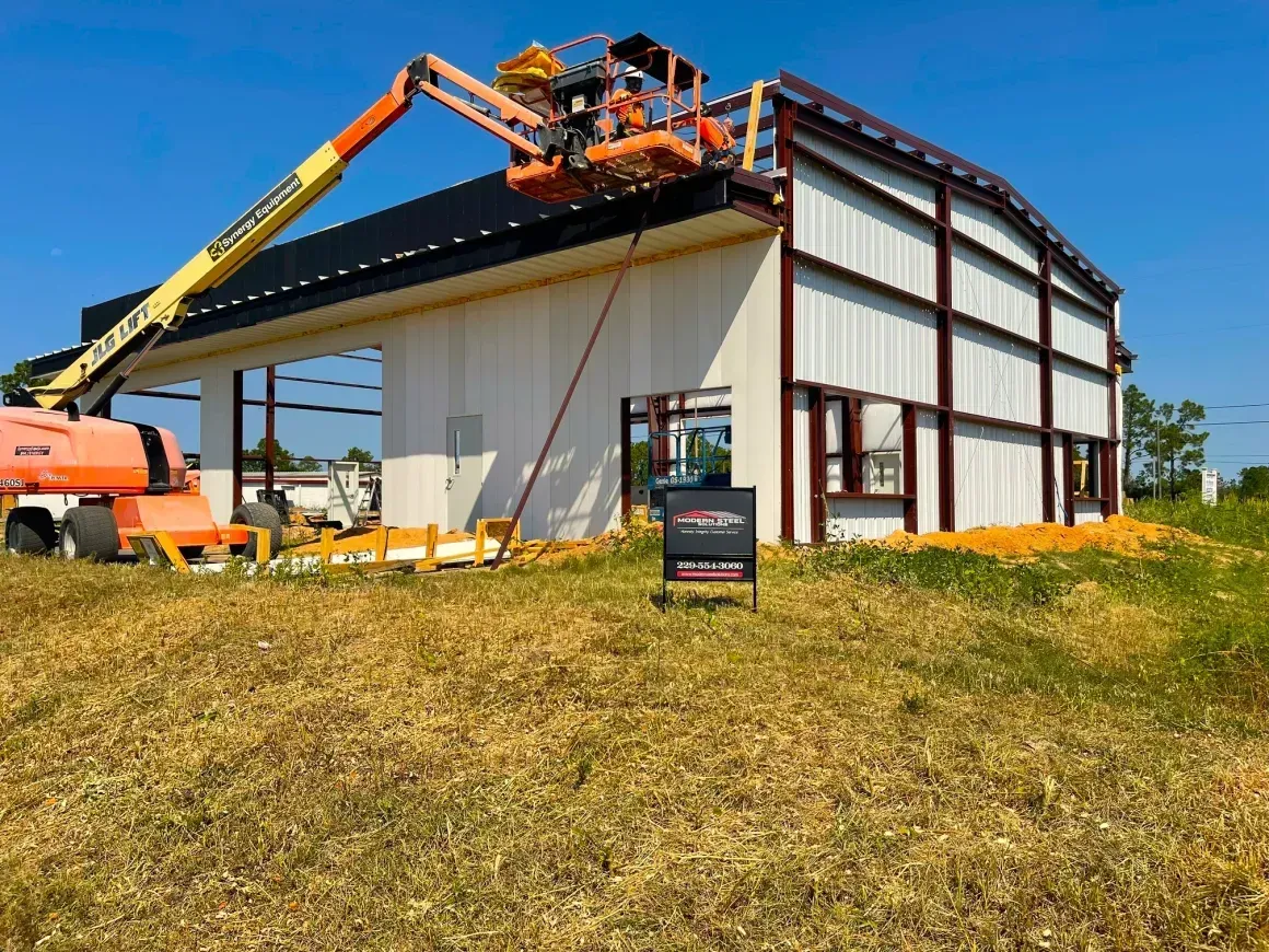 Construction workers using lifts on a large metal building with white walls and red trim under a blue sky
