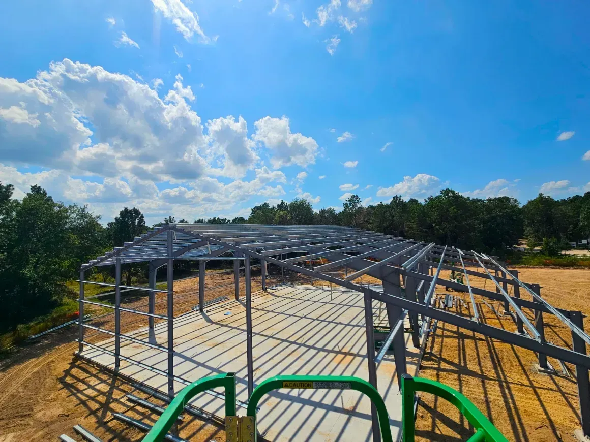 Steel building frame under construction on a dirt lot, viewed under a blue sky with clouds