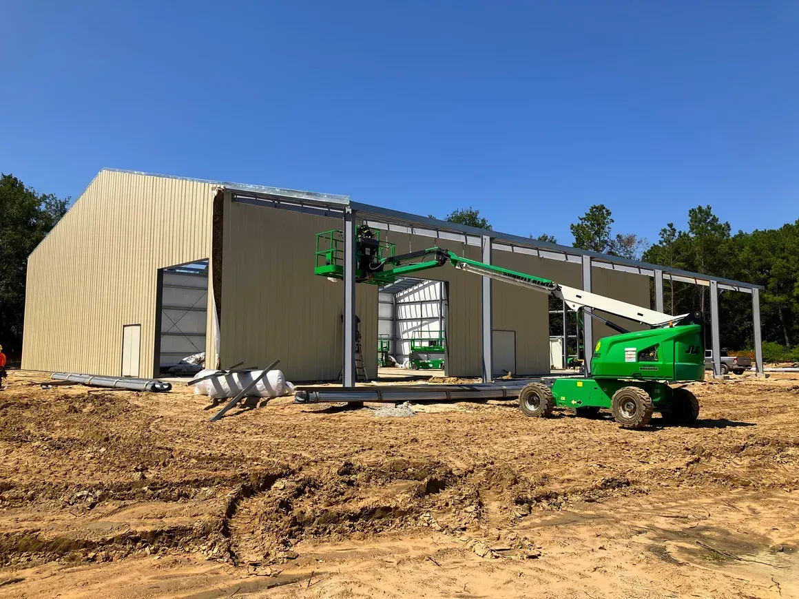 Construction site with a partially built tan metal building and a green lift on dirt under a blue sky