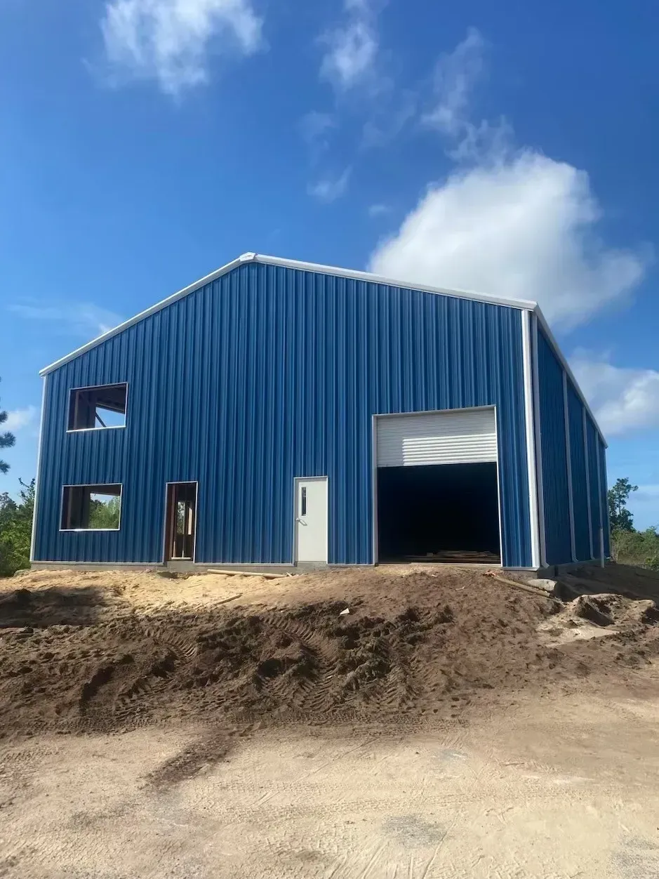 Blue metal barn under construction on a dirt slope beneath a partly cloudy sky